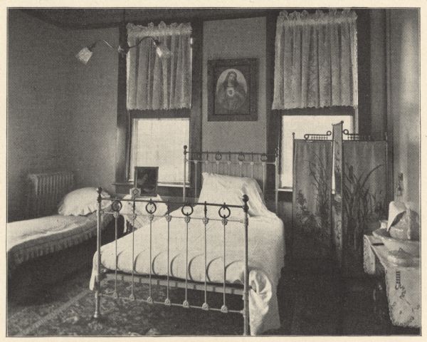 View of a large, private hospital room with a linen covered cast-iron and brass bed. Two windows with lace tier curtains allow light to enter the room, and a picture of the Sacred Heart of Mary is hanging on the wall between them. A daybed, steam heat radiator and bedside table holding a clock and framed picture are on the left. To the right is a three paneled folding privacy screen with a depiction of a nature scene.  Along the wall is a washstand with covered pitcher and basin. A large patterned floor rug and double-shaded hanging light fixture complete the view. Private rooms ranged from $10 to $20 per week, depending on the location and character of the room.