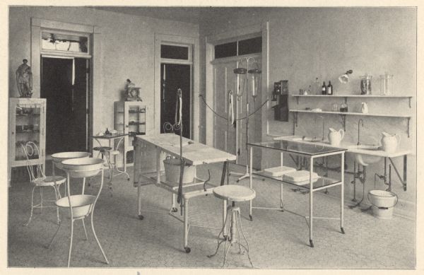 View of the brightly lit, tiled floor operating room. Cast-iron furniture includes an operating table, with a slanted central opening for drainage of fluids into a basin below. An I.V. stand with glass bottles is next to a wall-mounted telephone and wash basins. The following has been quoted from the hospital's 1902 patient booklet:  "The third floor is the surgical department. The operating room is on this floor, and special care has been exercised in its construction, looking to perfect arrangement for the latest and most approved methods in up-to-date surgery. It is lighted by a sky-light and large plate-glass windows. The floor is of vitrified tile with a base of the same material. Adjoining the operating rooms are the anesthetic and sterilizing rooms, and well equipped dressing rooms for the surgeons and nurses. This floor also has a well arranged room for the dressing of all surgical cases, and was constructed with a special view to surgical cleanliness. A number of pleasant private rooms occupy the remaining space on this floor." 