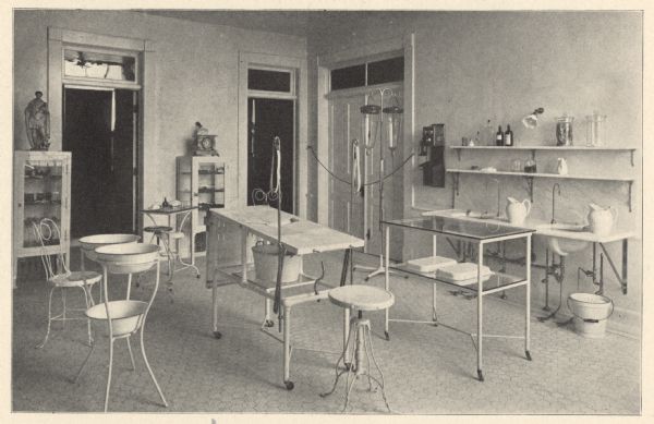 View of the brightly lit, tiled floor operating room. Cast-iron furniture includes an operating table, with a slanted central opening for drainage of fluids into a basin below. An I.V. stand with glass bottles is next to a wall-mounted telephone and wash basins. The following has been quoted from the hospital's 1902 patient booklet:  "The third floor is the surgical department. The operating room is on this floor, and special care has been exercised in its construction, looking to perfect arrangement for the latest and most approved methods in up-to-date surgery. It is lighted by a sky-light and large plate-glass windows. The floor is of vitrified tile with a base of the same material. Adjoining the operating rooms are the anesthetic and sterilizing rooms, and well equipped dressing rooms for the surgeons and nurses. This floor also has a well arranged room for the dressing of all surgical cases, and was constructed with a special view to surgical cleanliness. A number of pleasant private rooms occupy the remaining space on this floor."
