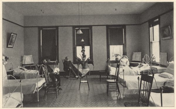 View of the men's first floor ward. Two bearded men are sitting on rocking chairs, dressed in street clothes and holding canes. A man is lying in a bed on the left, and a Sister is sitting beside the bed of another patient on the right. Furnishings include cast-iron beds, wooden spindle-back chairs and bedside stands. Large windows have dark shades and tiered lower curtains. Window sills hold plants, and a potted fern is on a linen-covered stand in the center of the room. Hanging from the ceiling are two electric lights with ruffled-edge opaque glass shades. Hardwood flooring appears to have a central parquet design. Ward patients were charged $1 per day, which covered board and nursing, only. Charity patients were cared for free of charge.