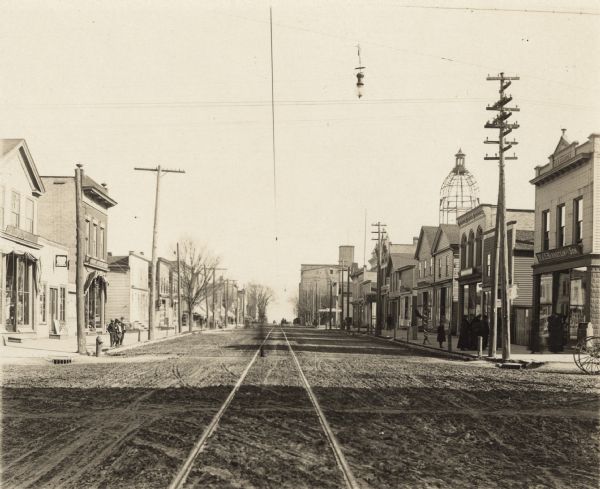 View of Washington Street from the intersection of South 10th Street looking east towards Lake Michigan. Streetcar tracks are running down the center of the unpaved street, towards the lake. On the right, women in dark clothing are gathered in front of Dr. J.E. Barnstein & Son's office and pharmacy at 925 Washington Street. Still under construction, the courthouse dome towers over the buildings. In the distance is the William Rahr Sons' large barley elevator. On the left, a group of boys are standing in front of Fred W. Meyer's hardware, tinware and paint store at 922-924 Washington Street. A lone electric street light is suspended over the intersection.