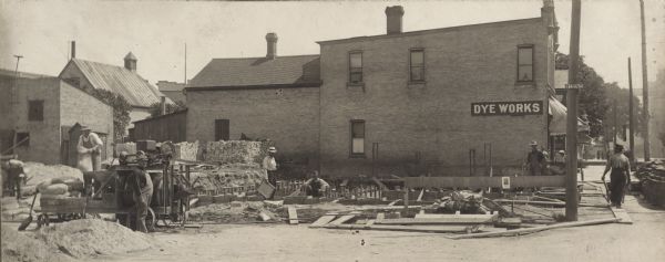 View of the construction site of the Glander Building on the northeast corner of S. 9th and Washington Streets. A masonry crew is constructing the foundation walls. On the left, men are working among sacks of lime, piles of sand and a mixer powered by a hit and miss engine.  Stacks of what are probably cinder blocks are in the background. On the right, another man is pushing a wheelbarrow, most likely filled with mortar, along a board to waiting workers. Henry Bode's large brick building, Manitowoc Steam Cleaning and Dying Works, is to the rear of the site. The Glander Building was the home of the Glander Art Studio from 1914-1972.