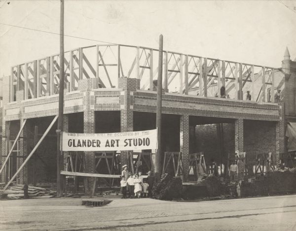 View across street towards a group of children standing in front of the Glander Building under construction on the northeast corner of S. 9th and Washington Street. A banner secured between two utility poles reads: "THIS BUILDING WILL BE OCCUPIED BY THE GLANDER ART STUDIO." Several workmen are laying bricks on the 2nd floor, while another man is looking out from behind a pile of bricks on street level. A narrow band of light-colored brick delineates the 1st from the 2nd floor. In the foreground, streetcar tracks are running down the center of brick-paved Washington Street.