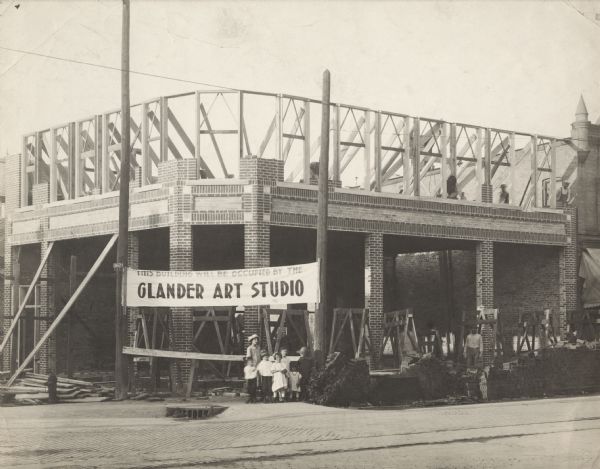 View across street towards a group of children standing in front of the Glander Building under construction on the northeast corner of S. 9th and Washington Street. A banner secured between two utility poles reads:  "THIS BUILDING WILL BE OCCUPIED BY THE GLANDER ART STUDIO." Several workmen are laying bricks on the 2nd floor, while another man is looking out from behind a pile of bricks on street level. A narrow band of light-colored brick delineates the 1st from the 2nd floor. In the foreground, streetcar tracks are running down the center of brick-paved Washington Street.   