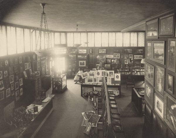 Elevated view of the 1st floor salesroom of the new Glander Art Studio at 822 Washington Street. Framed pictures are covering the walls. Tables and glass front cases are displaying prints, photographs, picture frame molding samples, frames, lamps and giftware. A woman is sitting near the corner entrance which is illuminated by daylight from the glass pane doorway and upper windows. A door on the rear wall gives access to the window display area behind it, facing S. 9th Street, while another door on the left allows entry to the Washington Street windows.  