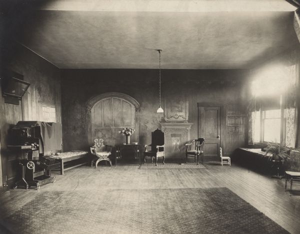 Daylight from a large, curtained window is illuminating the west end of the new photography studio. Furniture and props are arranged in front of walls with painted backdrop scenery. Rugs cover a hardwood floor, and a rolling studio camera holding a shelf of 8 x 10 film holders is on the left. The studio was on the 2nd floor of the Glander Building at 822 Washington Street.