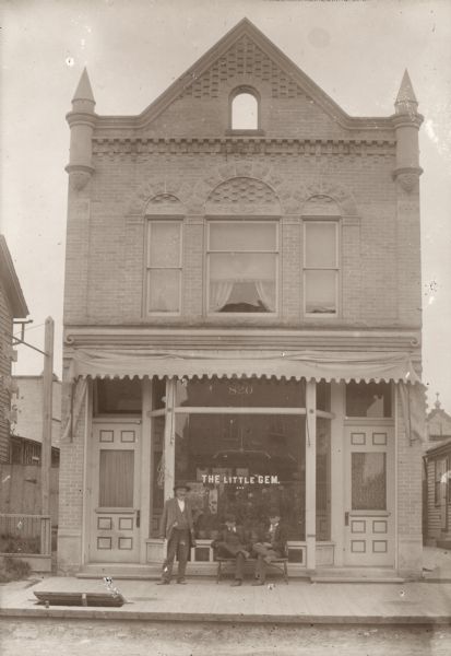 View of The Little Gem, a saloon and cafe at 820 Washington Street dating to 1895. Two men are sitting on a bench, while another man is standing next to them in front of the large first-floor windows.  A hatch door, partly propped open, leads to a space beneath the sidewalk and entrance to the basement.  The second-story of the brick structure features Romanesque Revival style Palladian arch windows, ornamental corner turrets and a corbeled cornice. Mathias Kettenhofen established his saloon and residence after selling the Northwestern House on Franklin Street, his well-known and highly-regarded hotel. An advertisement in a local German language newspaper in 1895 states that in the saloon of M. Kettenhofen, a fine glass of wine awaits the guest, Mosel wine above all, but also a large selection of other wines, the finest beers, liquors, non plus ultra cigars and everyday, a fine, good-tasting lunch. Many ads state the lunch is free. Mr. Kettenhofen retired in 1899, but The Little Gem. continued under other ownership until the property was sold in 1903 by William Rahr to Henry Bode for Manitowoc Steam Cleaning and Dying Works. John A. Glander acquired the building from Mr. Bode for the expansion of the Glander Art Studio, which was on the adjacent lot to the west, in 1922.