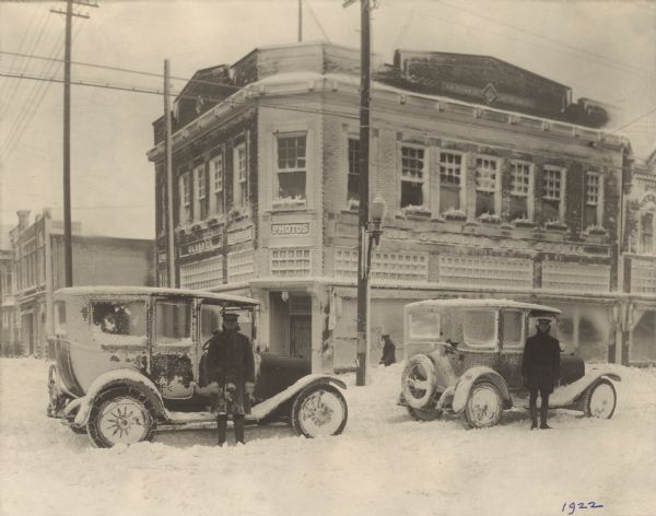 A covering of snow is clinging to buildings and vehicles on the corner of Washington and S. 9th Streets. Two men dressed in winter attire and professional-style caps are standing in front of cars with chains on the rear tires. Their occupations are unknown. Another man is looking on from the sidewalk in front of the Glander Building, home of the Glander Art Studio, at 822 Washington Street. A streetcar cable is suspended above the street.