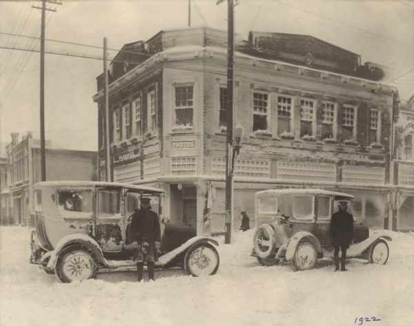 A covering of snow is clinging to buildings and vehicles on the corner of Washington and S. 9th Streets. Two men dressed in winter attire and professional-style caps are standing in front of cars with chains on the rear tires. Their occupations are unknown. Another man is looking on from the sidewalk in front of the Glander Building, home of the Glander Art Studio, at 822 Washington Street.  A streetcar cable is suspended above the street.