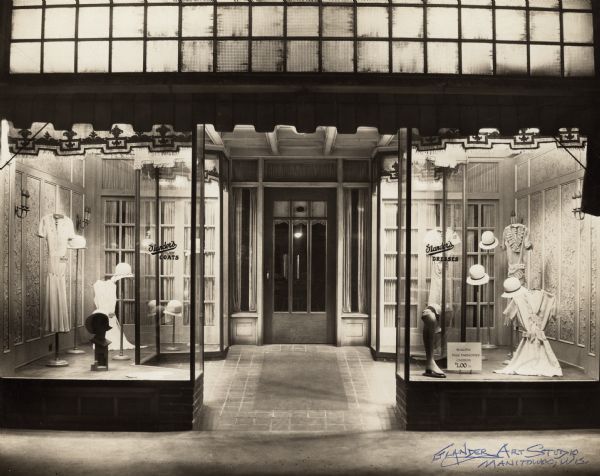 View of the Glander Style Shop's storefront entrance at 820 Washington Street featuring a twelve-feet-deep French window, tile floor, and heavy, ornate door. Women's summer dresses, hats and hosiery are on display. Window signs state: "Glander's Coats" and "Glander's Dresses." Newspaper ads offer ready-to-wear coats, dresses, hats, hosiery, brasiers, rayon underwear, nightgowns and pajamas for women and misses. John Glander purchased the building east of the Glander Art Studio for expansion of his business in 1922. It had been the site of Henry Bode's Manitowoc Steam Cleaning and Dying Works since 1903 and, for the last few years, Bode's Millinery Shop. After being remodeled, it opened as Glander's Hat Shop with John's sister, Hattie, who had prior millinery experience, as the proprietor of the shop. By 1927-'28 it was known as Glander's Style Shop. His brother William, was the proprietor, as well as being employed by John as a photographer and manager of the Glander Art Studio. The shop closed in 1931 so that both brothers could devote all of their attention to the increasing demands of the photography business. The shop space was, then, rented. The first occupant was Egan Apparel Shop in 1931. From the 1930's — early 1960's, other occupants were Pasewalk's, Smart Frocks, Jensen's and Houston's. All were women's apparel stores.