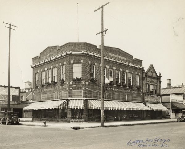 View across intersection towards the Glander Art Studio and the Glander Style Shop on the northeast corner of S. 9th and Washington Streets. Second-story windows are accented with flower boxes, and striped awnings are lowered above storefront windows. Built in 1914 to house John Glander's photography studio, art and giftware store, the Glander Building's original doors, one on the far-left corner and the main door on the canted, front corner, have been closed in with brick.  A new entrance has been constructed, facing Washington Street, midway between the store's display windows on the right. John Glander purchased the adjacent building at 820 Washington Street for expansion in 1922. The Romanesque Revival-style building, dating to 1895, was adjoined to the art studio and remodeled with a modern storefront and interior. It opened as the Glander Hat Shop with Hattie Glander, John's sister, proprietor. By 1928, it was known as the Glander Style Shop with John's brother, William, proprietor. William was also employed by John as a photographer and manager of the Glander Art Studio. On the far right, at 818 Washington Street, is Morris Alpert's shoe and men's furnishings store. On the left, a sign on the back of the two-story building advertises Henry Esch Sons Co. department store. The Schuette Bros. Co.'s water tank is in the distance.