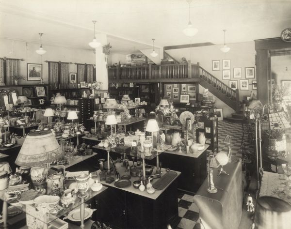 Elevated view of the interior of Glander Art Studio's gift shop, facing east. The wood framing of the entrance door from Washington Street is on the right. Giftware and lamps of all sizes are on display. On the far side of the room, a stairway ascends to an office balcony, overlooking the floor, and to the second floor photography studio. Founded in 1908 by John Glander, the business expanded to include original, imported fine-art pictures, reproductions, prints, custom-made and ready-made frames, mirrors, lamps, giftware, fine dinnerware and amateur photo finishing.   