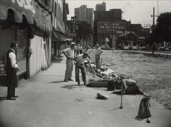 View down sidewalk along Washington Street facing east. Excavation is going on between S. 8th and S. 9th Streets. A crew is mixing concrete and doing work beneath the sidewalk. On the left, a man is standing in front of Jensen's, a women's apparel store, at 820 Washington Street. The Belle Shoppe, another women's store, is further down the block. Rahr Malting Co. and the Colonial Inn are in the background. Before repaving could begin, underground work included lowering and enlarging sewer and water mains, putting in laterals, gas pipes, conduits for new street and traffic lights and rebuilding the sidewalk basements where they would project into the widened street. Old streetcar rails, extending east from the 13th Street intersection, had been covered up for many years. They were torn up and sawed into lengths to be taken to junk yards. The old paving bricks that weren't broken by the power shovel were carted off to be saved for the city's future building needs. The Washington Street improvement project ran from S. 8th to S. 15th Street, replacing the old brick and asphalt surface with a concrete street slab. The previous 50 foot width was increased to 56 feet by removing 3 feet of sidewalk width from both sides of the street. Widening allowed for four lanes of traffic plus room for parallel parking on each side. The City of Manitowoc paid for the preparations and finishing work; new walks and entrances to properties. The $150,000 paving job was paid for out of state and federal funds because Washington Street was part of a connecting link between state and federal highways through the city. The original plan included S. 7th to S. 8th, as well. However, Hwy. 10 was rerouted south on S. 8th from the Washington Street intersection. The block was not eligible for funding and was not repaved.