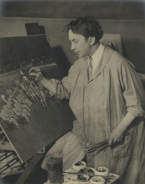 Three-quarter length portrait of John A. Glander in his artist's smock retouching a large photograph of a band. Mr. Glander was, for the most part, a self-made artist photographer in his mastery of the use of camera and lighting, processing negative films, printing positive images and retouching of films and prints. As a color artist, he retouched portraits with some oil paint but preferred water color. He studied paintings and artists with great interest and, in his early years, palmistry was an avocation. He was especially concerned with posing hands gracefully in portraiture. When not occupied with photography, music and art were primary interests.