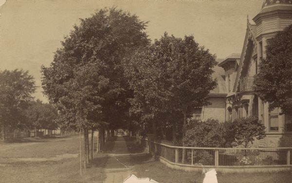 View down tree-lined sidewalk along Marshall Street facing west.  The northwest corner of S. 6th and Marshall Streets is circled by a fence, which continues down the block in front of two homes. Grass lines the walk and extends into the unpaved street. The Queen Anne style home at (202) 602 Marshall Street, with its corner tower and gingerbread trim, is the residence of Louis Pitz, City Engineer. The adjacent residence, obscured by trees, at (206) 606 Marshall Street would later be moved to 1210 S. Lake Street. The brick entrance tower of the High Victorian style home has a square, corbelled roof. A glimpse of the front bay window features a narrow Gothic arched window with upper trefoil pattern and surrounding rope-twist trim. Built ca. 1875, it was the homestead of August F. Dumke until 1890. Mr. Dumke (1824-1906) was a native of Germany who arrived in Manitowoc County in 1853. He served in the Civil War as 1st Lieutenant of Company B, 9th Wisconsin Infantry and was promoted to Captain in less than a year. He owned and operated Manitowoc Iron Works on Quay Street.  His foundry and machine shop manufactured steam engines and provided machinery for early mills and other industrial needs. The residence was acquired in 1912 by William Rahr, II (1854-1919), whose adjacent home was at 612 Marshall Street. According to information passed down through the Rahr/Spindler family, the home was moved from 606 Marshall Street to 1210 S. Lake Street as a wedding gift to his daughter, Martha Rahr, when she married Walter E. Spindler in 1914. In 1963, the home was donated to the Manitou Girl Scout Council as a memorial to Martha Rahr Spindler (Mrs. Walter E. Spindler), a founder of the Girl Scout movement in Manitowoc.