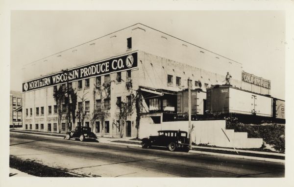 View across street towards the warehouse and cold storage building of the Northern Wisconsin Produce Company at 1310 Clark Street. Two workmen are icing the freight cars. Ice is made in 300 pound blocks in the ice house on the second floor of the cold storage at the far end of the facility. Using long, hooked poles, the workmen slide the blocks along the shipping dock roof onto the cars, dropping them into spaces on both ends. Before mechanical refrigeration, ice was used to keep cheese and produce cold during shipping. The Northern Wisconsin Produce Company was incorporated in 1907 by Stanley Eckels, Charles E. Spindler and Gustave Lindemann. They purchased the cold storage facility of the Northern Grain Company on the northeast corner of S. 6th and Quay Streets, of which Stanley Eckels had been the manager.  Mr. Eckels became the head of the new company and manager, when it opened in 1908. The new plant, opening in 1925, was built on the site known as Truman's Hill at S. 14th and Clark Streets at a cost exceeding $100,000. The hill was cut down, and a modern, brick cold storage and produce shipping plant was built by Schuette Construction Company. A spur track entered the grounds from the adjacent Chicago & North Western Rail Road's main lines. The firm was a wholesaler of cheese, butter, margarine, eggs, and other farm products.  At this time, the officers of the company were Stanley D. Eckels, Victor Trastek and Gustave Lindemann. The Northern Wisconsin Produce Company is owned and operated by fourth generation members of the Lindemann family as a cheese and cold storage business, as of 2018.