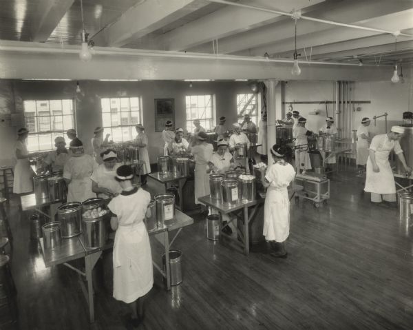 Long white gowns cover the street clothing of women at work separating egg yolks and whites into glass cups in the egg-breaking room of the Northern Wisconsin Produce Company.  Metal canisters, some filled with fresh, whole eggs and others holding the separated eggs, are lined up on each work table.  Underneath, large containers catch broken shells, which are dropped through holes in the center of the tables.  A man in a white apron and cap is carrying a canister and another man, on the right, has his hand on the spigot of a churn.  Sanitary, hospital-like conditions of cleanliness are being closely maintained.  Located at 1310 Clark Street, the breaking room is on the second floor of the building, facing west.  Eggs coming into an egg-breaking plant were first candled to grade for quality.  Those that passed certain conditions of freshness went to the breaking room to be separated into yolks and whites, although some were processed without separating.  The canisters of yolks and whites were emptied into churns, mixed, drawn off through spigots into cans, commonly holding 25 pounds, and sent to cold storage for freezing.  Room temperature was regulated in each of the processes and equipment was sterilized, frequently.  The Northern Wisconsin Produce Company was a wholesaler of eggs, cheese, butter, margarine and farm products, which it purchased from Wisconsin farms and factories.    