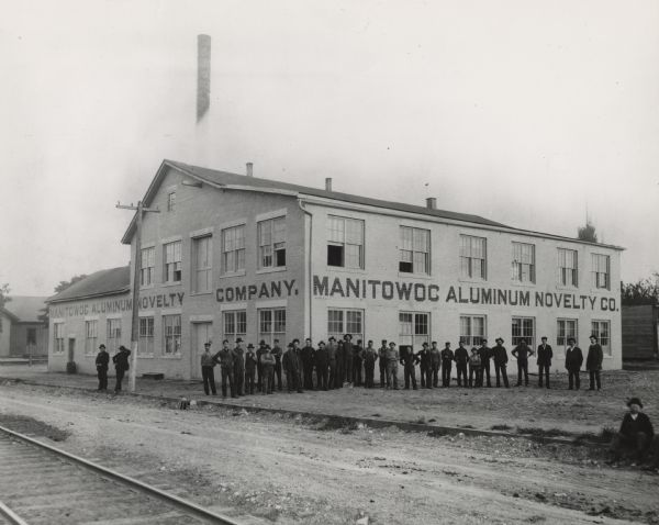 View across Franklin Street of the Manitowoc Aluminum Novelty Company on the southwest corner of S. 15th and Franklin Streets. This very first picture of the plant, which is facing the north and west sides of the building, reflects the humble beginnings from which the Mirro Aluminum Company grew.  An article in the "Manitowoc Herald News" identifies the men, from left to right:  William Wellert and Peter Kaufmann, at the extreme left, are not employees of the company.  The others, in order, are Julius Schrank, Adolph Pelishek, Herman Schwab, Fred Lippert, August Willert, John Brobka, Alfred Schwab, Lawrence Zawatske, Alfred "Bud" Behringer, Charles Singleman, A. Todl, John Pelishek, Henry Meihsner, Herman Pentzien, Charles Nack, Jr., August Daugs, Fred Heise, John Mosuch, William Schaefer, Adam Specht, Robert Wiesman, Otto Manthe, Ferdinand Veith, James Lee, Max Auman, Charles Nack, Sr., Henry Vits (first president and treasurer), George Vits (future president), William Pflueger (first secretary), and a salesman named McMahon who was a successful seller, that day, and remained long enough to be included in the picture.  The person in the lower, right corner is not identified.  The company occupies a former tannery built in 1866.  Henry Vits (1842-1921) became associated with the tannery of Martin Vollendorf on S. 15th and Franklin Streets in 1873.  The firm is listed as "Vollendorf & Vits" in the 1875-'76 city directory.  In 1881, he became its sole owner.  The 1893 Columbian Exposition in Chicago introduced novelties imported from Germany which were made of aluminum, a new type of white metal.  At a time when the leather industry was changing and inspired by possibilities of a profitable merchandise line, Mr. Vits re-equipped the tannery in 1898 to produce aluminum combs, advertising novelties and other small products.  A 1909 merger with the Aluminum Manufacturing Company of Two Rivers, Wisconsin and the New Jersey Aluminum Company of Newark, New Jersey resulted in the name being changed to the Aluminum Goods Manufacturing Company.  Several generations of the Vits family continued to direct operations.  In 1957, the name was changed to the Mirro Aluminum Company.  It was known as the world's largest manufacturer of aluminum cookware.