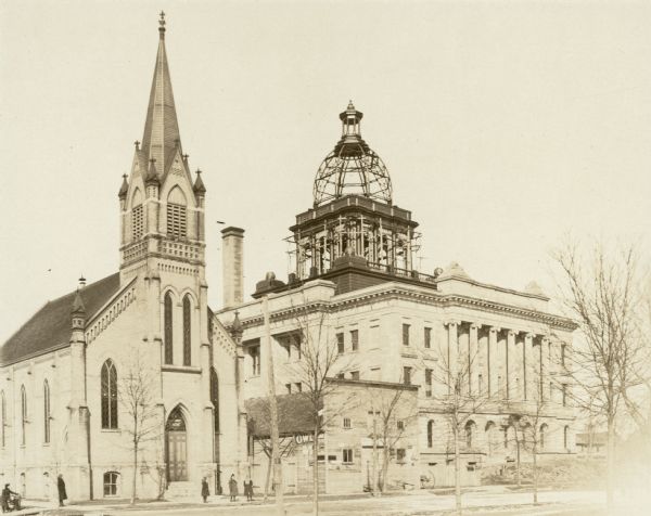 View facing north east across Hancock Street towards the Evangelical Reformed Church, also known as the German Reformed Church, and the new Manitowoc County Courthouse. Several girls wearing long coats are standing on the sidewalk in front of the church. Architectural details include tall, narrow Gothic arched windows, corbeled gable end and ornamental turrets. The entryway displays "Ev. Reformirte Kirche" in large letters above the double doors. Erected in 1891 on the corner of S. 9th and Hancock Streets, the brick church cost in excess of $6,000. The Beaux-Arts style courthouse, designed by Manitowoc architect C.H. Tegen and built of Indiana limestone, is in its final phases of construction. The framework of the dome and its base will soon be finished in copper with prism glass windows furnished and installed by the Tremmel Art Glass Company of Two Rivers, Wisconsin. At a cost of approximately $250,000, the courthouse was dedicated on Nov. 12, 1907.  One of the featured speakers, that day, was Wisconsin Governor James O. Davidson. A livery stable constructed of wood stands between the church and courthouse. A partially obscured sign on its left side advertises "OWL [CI]GAR 5 cts."