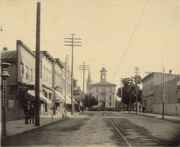 "About four o'clock I took a snapshot of the courthouse, which is now removed as far as on eighth Str.," wrote photographer John A. Glander in his diary entry of July 14, 1905. Two men are standing in front of the courthouse entrance and others are observing from the sidewalks.  Glander's view of the 1856 three-story brick structure captures its move from the courthouse square onto S. 8th Street, between Washington and Marshall Streets. It's being moved to 1026 S. 8th Street, the lot south of the jail, to make room for construction of a new county courthouse. Henry Buestrin & Sons of Milwaukee has been contracted to do the work at the price of $3,960. Several layers of hardwood-beam racking, each layer turned 90 degrees, support the building's extreme weight. It's likely that long, debarked and greased hardwood logs are being used beneath the racking. As oxen pull the load forward, in this case away from the viewer, the building is slowly rolled over the logs to its destination. In the foreground, signs and businesses between Jay and Washington Streets include an apothecary post for C.A. Groffman's drug store, Doctor Huwatchek, Dentist, and A.P. Schenian, Lawyer. All three are in the Williams Block, which is out of view to the left.  "Hotel" marks the German House awning, and the hardware store of Wernecke & Schmitz is on the corner.  The two-story brick building and fenced yard on the right is Fred Lohe's meat market.  Streetcar tracks run down the center of the street.