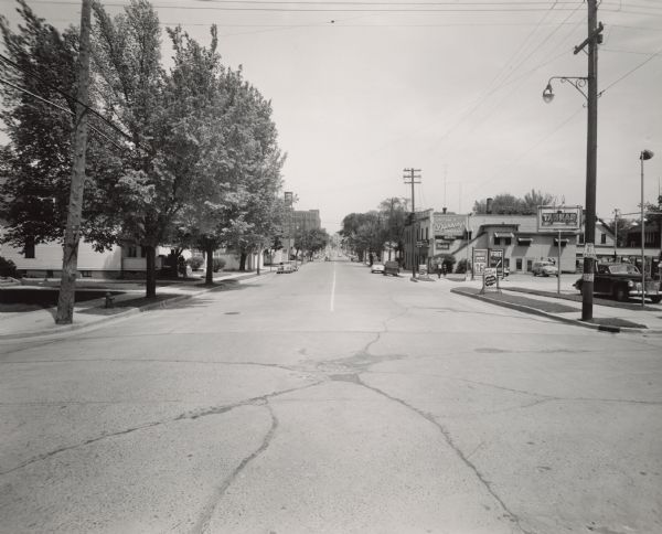 View of Washington Street from the intersection of South 19th Street, facing east. On the right, a sign identifies the corner location of the Tankar Gas Station. Additional signs advertise gas at 26.9 cents per gallon with premiums of free tableware. Beyond the station, a sign on the two-story building states "Stop and Shop at Dunning's Grocery." An enclosed exterior staircase leads to the second floor apartment. The store's Chevy panel delivery truck is parked on the side of the building. On the distant left, a rooftop water tower marked "MIRRO" identifies the Aluminum Goods Manufacturing Company. Several residences and businesses are partially obscured by trees.