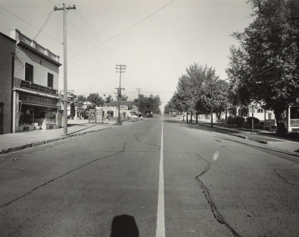View of Washington Street from South 18th Street, facing west.  Signage identifies the two-story structure on the left as Dunning's Clover Farm Store. Patterned tiles surround the storefront windows, and a "Kingsbury Beer" sign is hanging from the facade. The adjacent Tankar Gas Station, managed by Merlin Rupelt, offers gas at 26.9 cents per gallon. Dufek's Sinclair Service Station is on the next corner.  On the right, a highway marker denotes US 141, 151 and WIS 42.  Dunning's, at 1805 Washington Street, was a grocery, liquor store and bakery owned by Edward C. and Margaret Adams Dunning.  It was co-managed by Harold R. Monka and Christian (Ket) Peitersen, who were also the store's butchers. Mr. Peitersen's wife, Adeline, was the bookkeeper, and the family lived above the store. Boys of high school age were employed as stock boys and girls as clerks after school and on weekends. Dunning's was not a self-service grocery; the customer called and gave a list or told a clerk at the counter what he or she wanted. The clerk gathered the items, and the purchase was completed.  Delivery to customer's homes was done, as well. The store's popularity was partly due to its outstanding homemade bakery and delicatessen items prepared from Mrs. Dunning's recipes and the fact that it was one of the very few stores open on Sunday. An after-church stop and wait in line beneath the light green-colored pressed tin ceiling for hot, sliced ham, fresh bread and hard rolls, baked beans, German potato salad, meat loaf and other specialties was a cherished tradition for many families. Previously known as The Adams Bakery, the property was acquired in 1914 by Edward and Margaret Dunning from her father, Thomas J. Adams. It was in operation until 1970.