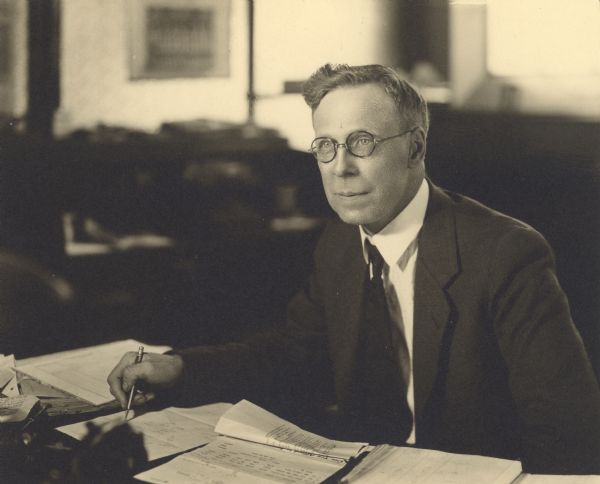 Portrait of Ralph Gordon Plumb (1881-1976), president of the Plumb & Nelson Company, seated at his desk. The company was a wholesale grocery business at 718 Buffalo Street. It was established as a retail business by his father under the name J.E. Plumb & Bro. in 1873. John E. Plumb and his younger, half-brother Julius C. Nelson were dealers in groceries and provisions, flour and feed, according to early newspaper advertisements. By 1893 the firm had been changed from a retail to a wholesale enterprise. After graduating from the University of Wisconsin in 1901, Ralph joined the family business. He became its president in 1923. During his college years in Madison, he spent much of his free time at the State Historical Society Library accumulating data from archived newspapers covering fifty years of Manitowoc's early growth. He published "A History of Manitowoc County" in 1904, "Born in the Eighties" in 1940 and several additional works of local, state and national history. He was very involved in civic affairs and organizations. In 1922, he was a founding member and the first president of the Manitowoc Kiwanis club.
