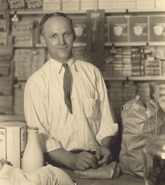 Waist-up portrait of Bernard H. Mahnke (1891-1978) standing behind a counter packaging groceries, possibly for delivery. Merchandise lines shelves in the background. Mr. Mahnke owned and operated Mahnke & Multerer, 1302 Washington Street, from 1918 to 1960 along with Joseph Multerer, Jr. The neighborhood grocery store was a member of the Manitowoc Retail Grocers' Association of which Mahnke was a charter member and president. It became a Clover Farm store in 1933 with the opening of the Eastern Wisconsin Division of the Clover Farm Stores system. The Plumb & Nelson Company was the local wholesale supplier.  Newspaper advertisements in 1933 offered items such as freshly-ground coffee at $.25 per pound, flour at $.71 for a 24 1/2 pound bag, three pounds of fresh tomatoes for $.25, four bars of Palmolive soap for $.25, and a large bunch of California carrots for $.05.  
