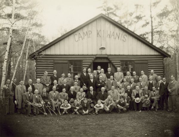 Group portrait of Kiwanis Club members and several boys assembled in front of the Camp Kiwanis cabin in Lincoln Park. The boys may be part of Boy Scout Troop 12, which was sponsored by the club. The Manitowoc Boy Scout Council had its summer camp, Camp Sinawa, at Pigeon Lake.  Camp Kiwanis, in Lincoln Park was its winter camp. Girl Scouts and other environmental and civic groups also made use of the facilities. Kiwanis Club members originally constructed the log building in 1923, mainly with their own labor, on city-owned property just north of what is now Reed Avenue and just west of the Little Manitowoc River. The building, which was 24 x 40 feet, had 16 double-bunks, cookstove, heating stove and a rustic fireplace constructed with colored field stones from the nearby, city gravel pits. The river wound its way through the flats one hundred feet to the east of the camp. The site was ideal for work in forestry, skating and skiing. Each troop reserved the shack, as it was called, for one or more weekends from fall through spring for overnight camping and scout activities.  The cabin was moved to Lincoln Park late in 1932 because it was frequently damaged by hoodlums and used by hobos as a place to sleep.