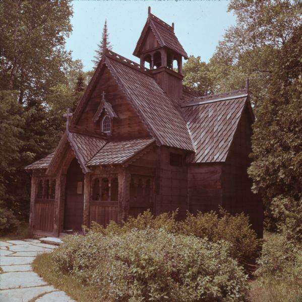 A wooden chapel with a stone walkway sitting among trees and bushes. Crosses are above the door and above a small window. A bell is hanging in the bell tower. Decorative patterns adorn the eaves and ridges of the roof.
