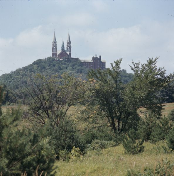 View across fields and trees towards the Holy Hill National Shrine of Mary on the top of a tree-covered hill.