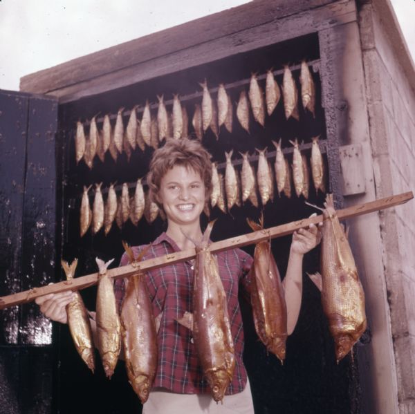 A woman is standing outdoor in front of a smokehouse and holding up a wooden rod with smoked fish hanging from it. More fish are hanging from rods behind her in the smokehouse.