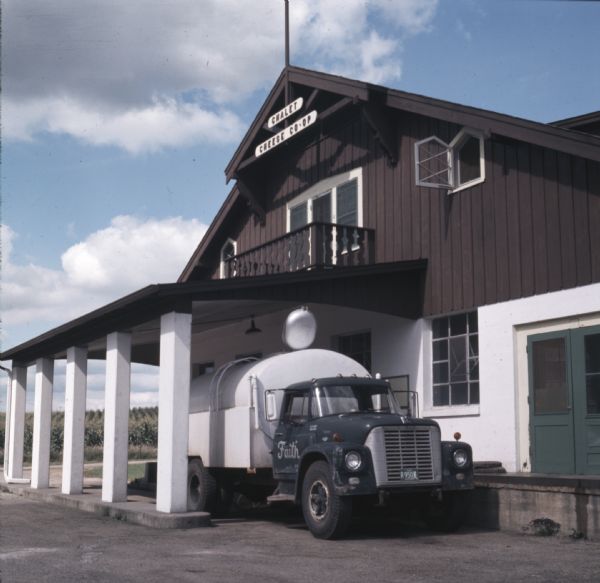 A dairy truck is under an awning at a cheese factory. A sign near the top of the two-story building reads: "Chalet Cheese Co-Op." Field crops are growing behind the factory.