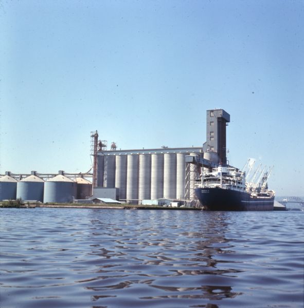 View across water towards a cargo ship at dock in front of large grain elevators.