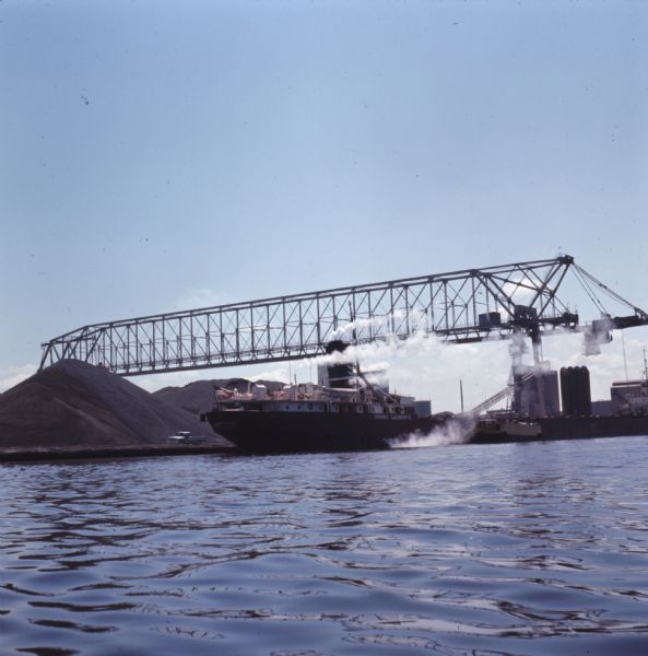 View across water towards the cargo ship "Henry LaLiberte" sitting in a loading dock under a cargo crane. 