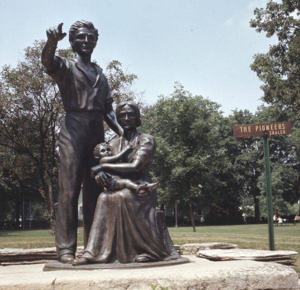 Bronze statue of a standing man pointing off into the distance, with his wife sitting beside him holding a baby in her lap. The statue sits on a stone platform. A small sign behind the statue reads: "The Pioneers, Shaler."