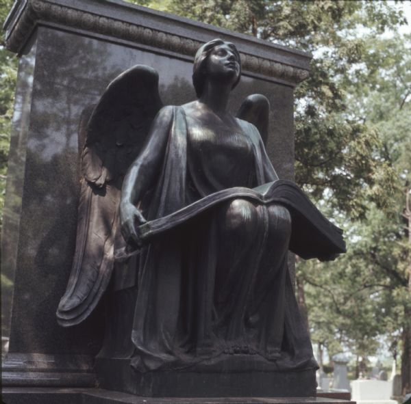 A bronze statue of a female angel holding a large book in her lap is in front of a marble gravestone. Gravestones are in the background on the right.