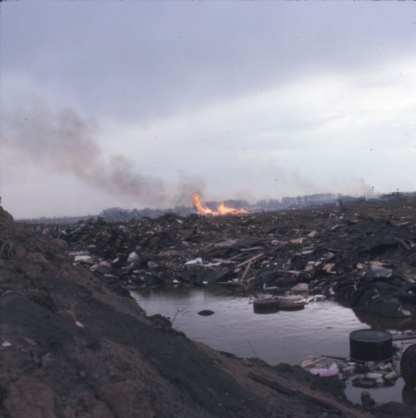 View across garbage and waste around a pool of water towards a fire burning in a landfill. Trees are along a ridge in the far background.