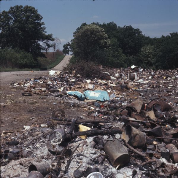 Solid waste, some of it burned, scattered on the ground in a landfill next to a road along the left side. In the background are trees and a hill.