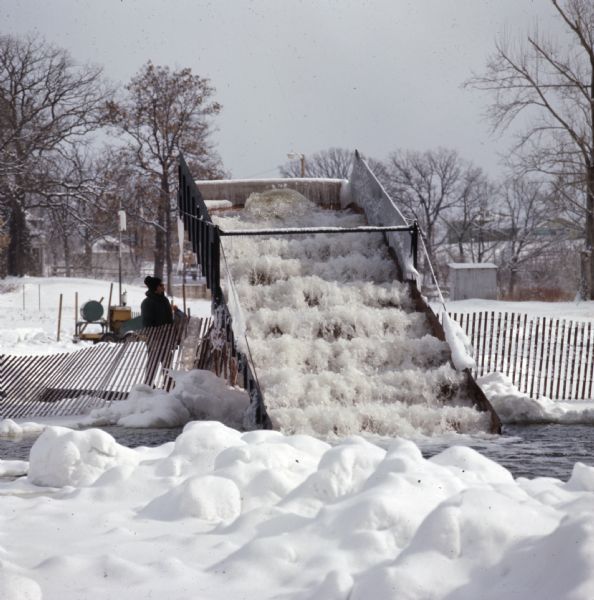 A man is standing behind a snow fence next to a surface aerator. Water is pouring down or up the machine, breaking the surface tension of the lake. Snow is on the ground.
