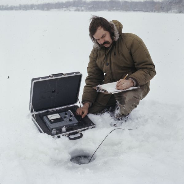 A man is kneeling on the ice and snow at Potter's Lake. He is running a machine that is testing the oxygen levels in the water, while recording the results on a clipboard.