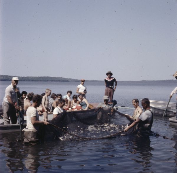 View across water towards men, women, and children sitting in metal boats on a lake. They are watching five men standing waist deep in the lake holding up a seine net filled with fish. 