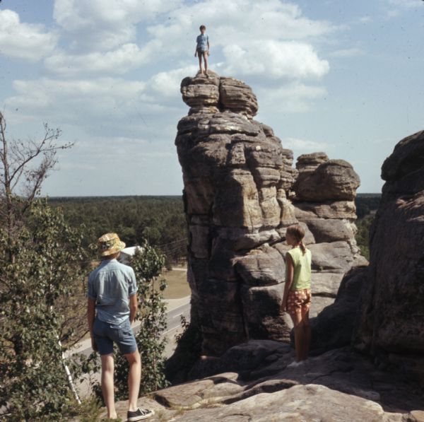 Three young people are standing on Rabbit Rock, two of them in the foreground, and one on top of the taller peak. Highway 13 is below in the background.