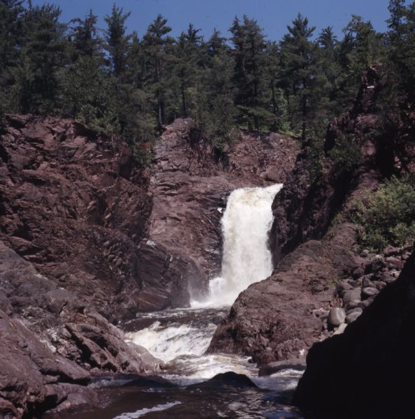 View of Brownstone Falls on Bad River. Rock formations are on the left and right banks. Trees are along the top of the rock formations.