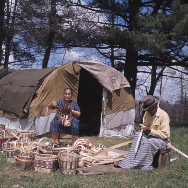 Mable and Martin Lowe sitting outdoors, with a dwelling (chipoteke) behind them. Mable is weaving a basket, while Martin is working on strips of wood for handles. Several finished baskets are on the ground in front of them.