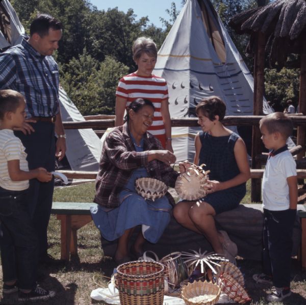 Sarah Snake (WhiteEagle) is sitting on a bench and showing a tourist how to weave a basket. Other visitors are standing around her to watch the demonstration. Strips for weaving and finished baskets are on the ground in front of the bench. Two tepees or tipis are in the background.