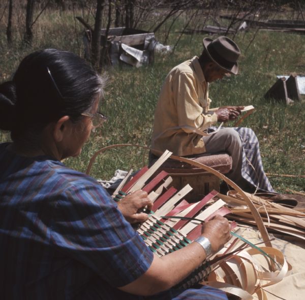Over the shoulder view from behind of Mable Davis (maiden name) Lowe as she is weaving a basket outdoors. Martin Lowe is sitting nearby on a cushioned bench preparing handles.