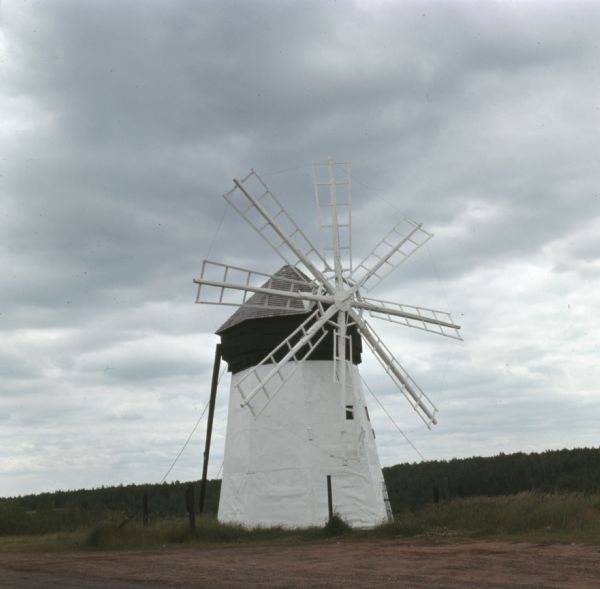 View of a white and green windmill with eight blades is surrounded by a small fence in a rural area. Trees are in the far background.