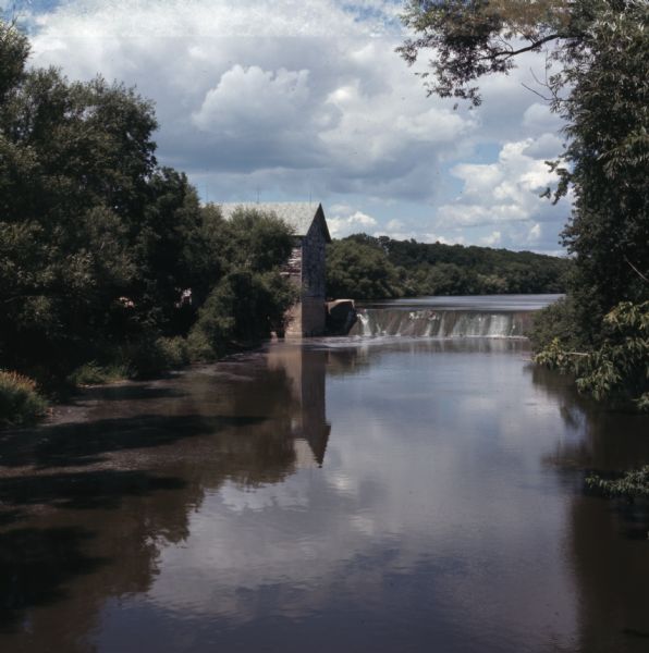 View looking upriver towards the Jaeger Rye Mill, also known as the Danville Mill, on the left in front of a small dam. Trees are along the banks of the Crawfish River. The lower half of the mill is built of stone, and the upper stories are wood. 