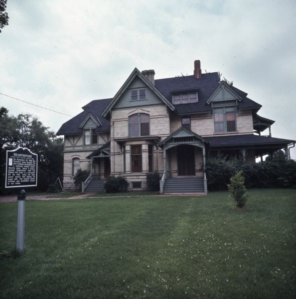 View across lawn towards the historic Hearthstone House. A historic marker is in the foreground on the left, and it reads: "Hearthstone, World's first Electric Lighted Home. On September 30, 1882 this Victorian home became the first residence electrified from a centrally located hydro-electric plant. Henry J. Rodgers, pioneer industrialist, built and lighted this home as a showplace for his wife, using the same power source that lighted his paper mill. The original Edison switches and chandeliers remain in use today."
